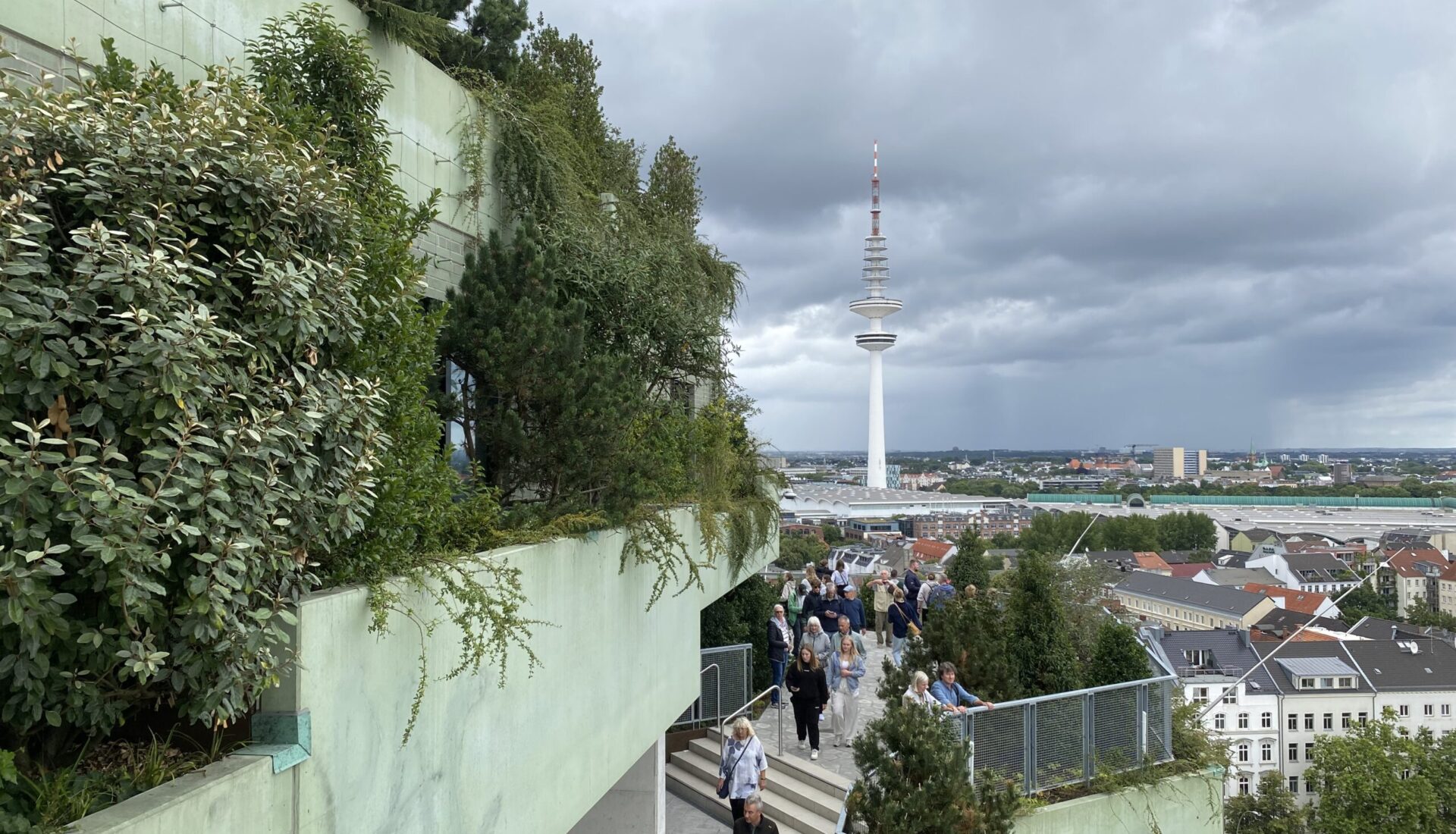 Menschen besuchenden Grünen Bunker in Hamburg, umgeben von Pflanzen und Bäumen, mit Blick auf den Hamburger Fernsehturm