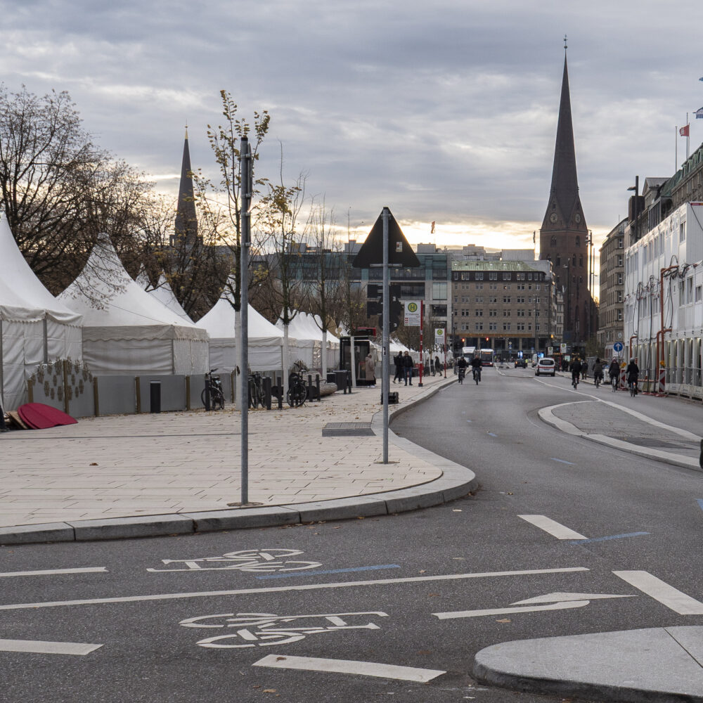 Der Jungfernstieg in Hamburg: Auf der linken Seite stehen die noch abgedeckten Stände des Weihnachtsmarktes, auf der rechten Seite ist ein Teil des Neuen Walls zu sehen. Hinten auf dem Bild erkennt man die Europa Passage und die Kirche.