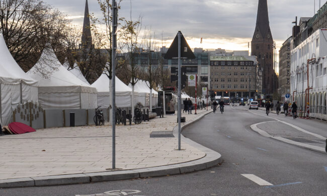 Der Jungfernstieg in Hamburg: Auf der linken Seite stehen die noch abgedeckten Stände des Weihnachtsmarktes, auf der rechten Seite ist ein Teil des Neuen Walls zu sehen. Hinten auf dem Bild erkennt man die Europa Passage und die Kirche.