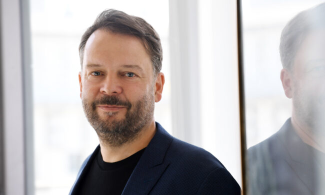 Ein Porträt von Michael Knobloch, dem Leiter der Verbraucherzentrale. Er hat einen Vollbart, braune Haare und blaue Augen. Auf dem Foto trägt er ein schwarzes T-Shirt mit einem dunkelblauen Jackett darüber.