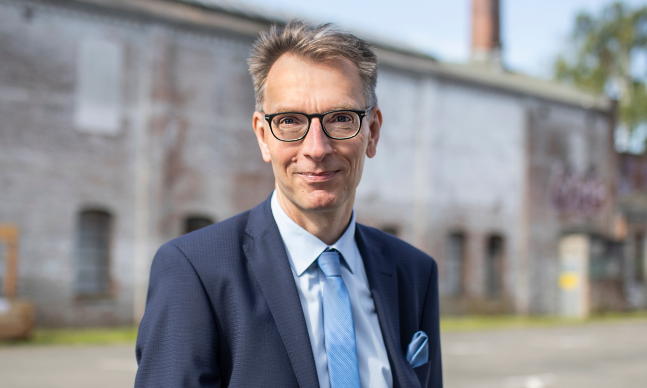 Mann mittleren Alters mit Brille und Anzug steht vor einem Gebäuderiegel.